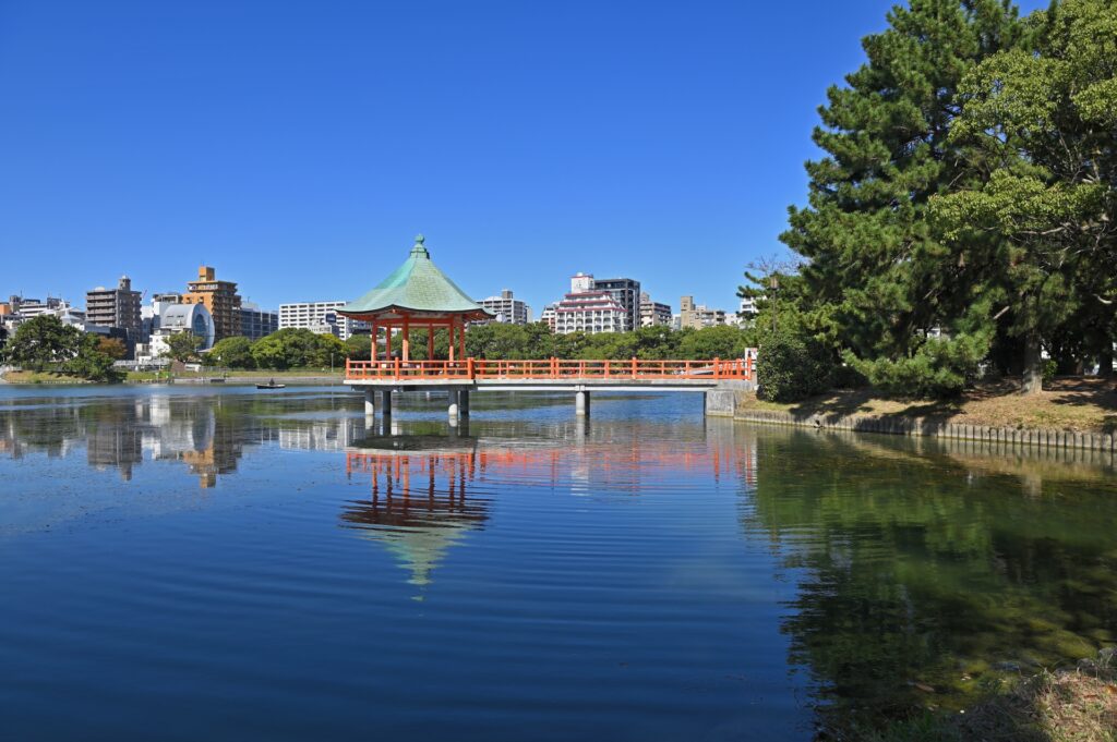 静かな湖面が広がる福岡県・大濠公園。水面には空の光が穏やかに映り、周囲の緑の木々が柔らかな風にそよいでいる。遊歩道には散歩を楽しむ人々の姿が見え、都市の中の大きな自然の安らぎを感じさせる風景。