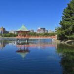 静かな湖面が広がる福岡県・大濠公園。水面には空の光が穏やかに映り、周囲の緑の木々が柔らかな風にそよいでいる。遊歩道には散歩を楽しむ人々の姿が見え、都市の中の大きな自然の安らぎを感じさせる風景。